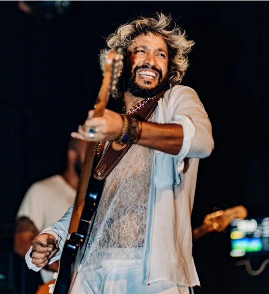 Em uma foto de plano médio, Leandro Léo é capturado durante uma apresentação musical no palco. Ele sorri amplamente, olhando ligeiramente para cima e para o lado, enquanto toca uma guitarra elétrica de cor escura. Seu cabelo está ondulado, com raízes escuras e pontas loiras descoloridas, e ele tem uma barba cheia. Ele veste uma camisa clara de botões, que está aberta, revelando por baixo uma blusa de manga comprida feita de renda branca e transparente. O fundo está escuro, com outro membro da banda e equipamentos de palco visíveis, mas desfocados.